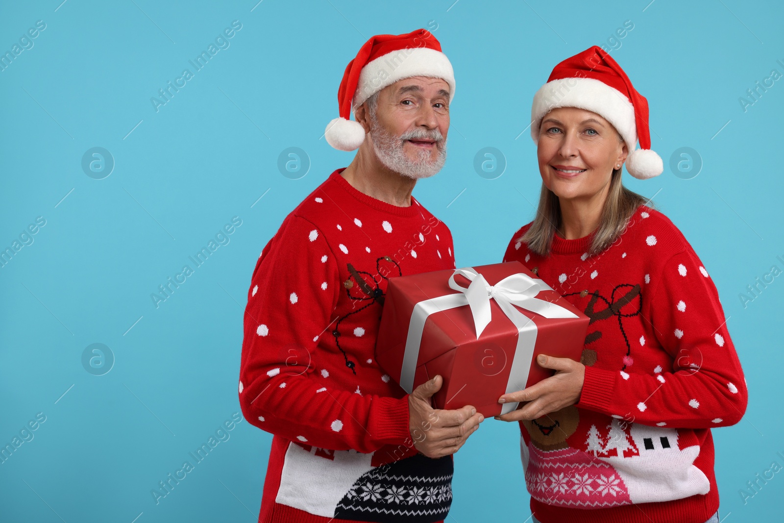 Photo of Senior couple in Christmas sweaters and Santa hats holding gift on light blue background. Space for text