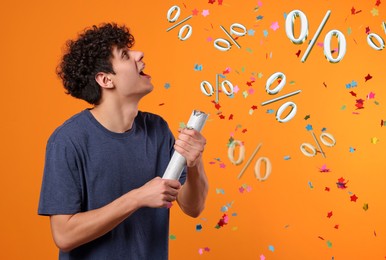 Discount offer. Happy young man blowing up party popper on orange background. Confetti and percent signs in air