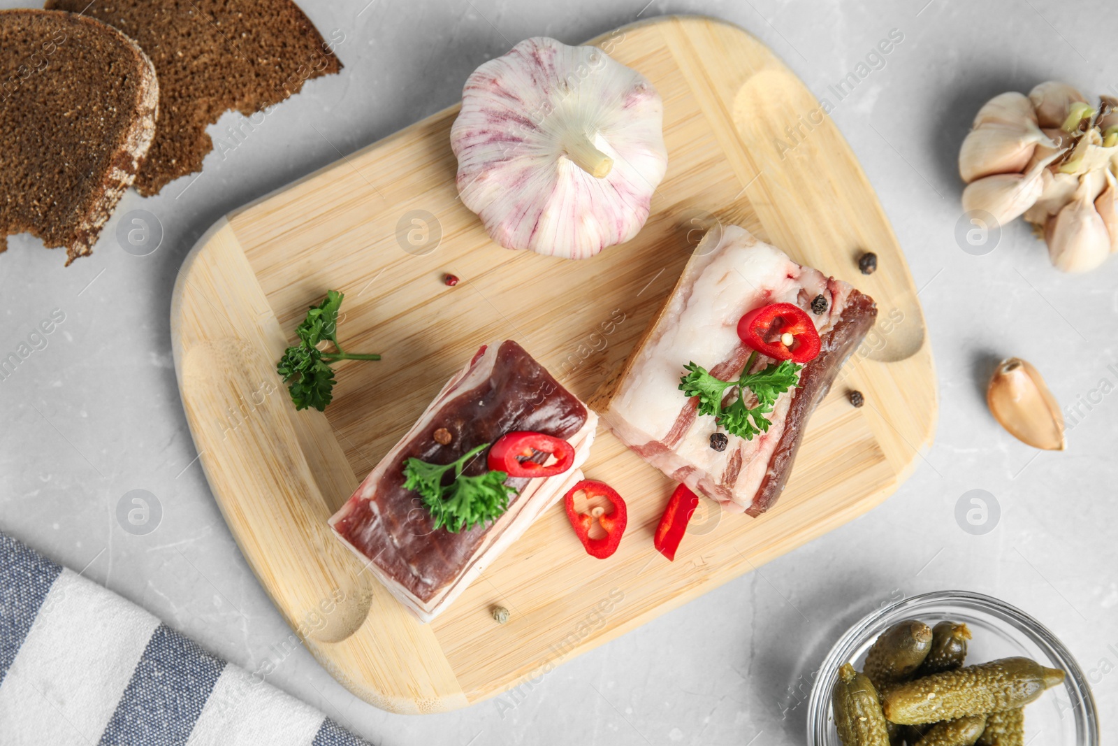 Photo of Board with pork fatback, rye bread, garlic and bowl of pickles on stone background, flat lay