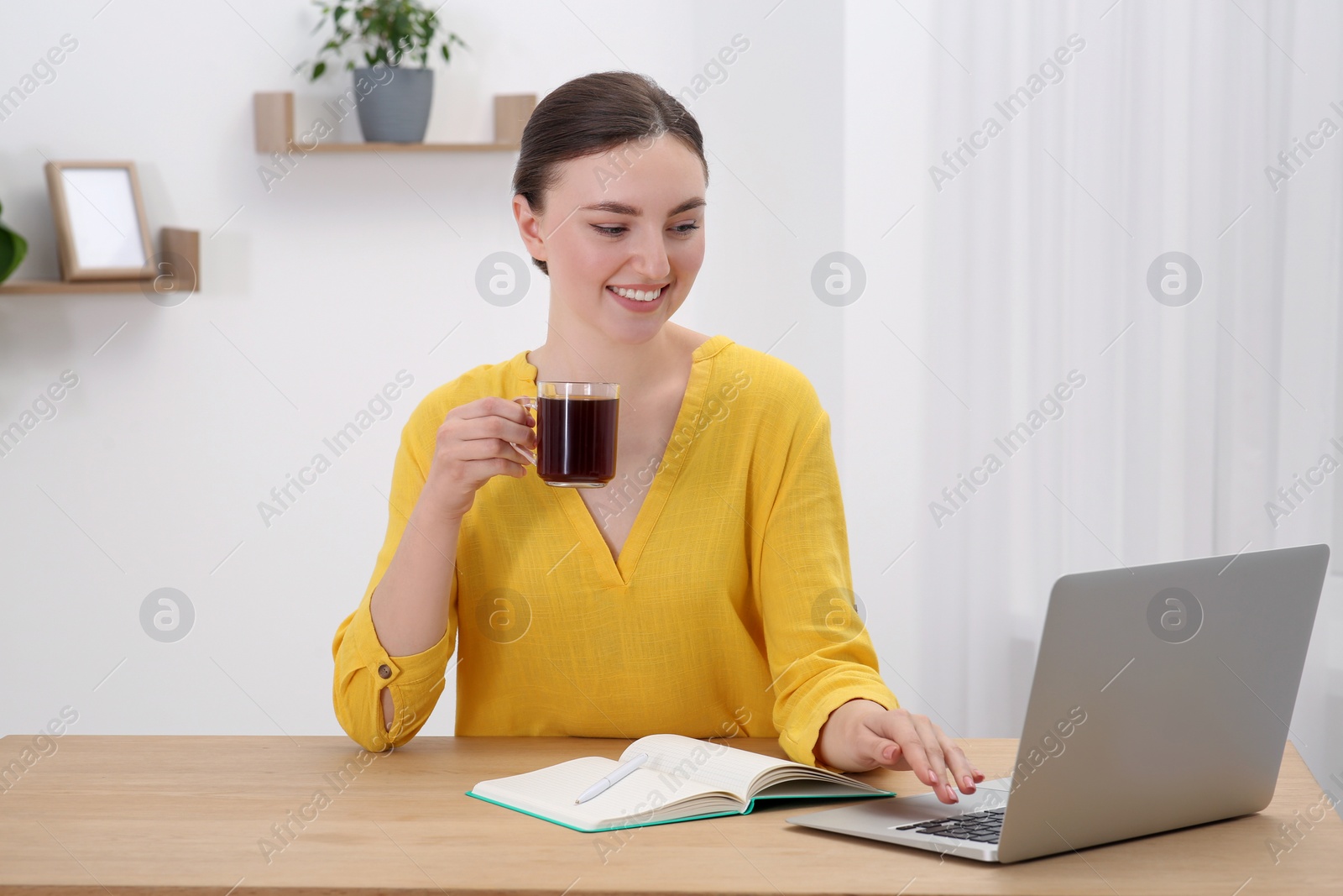 Photo of Happy young woman with cup of coffee and notebook working on laptop at wooden table indoors