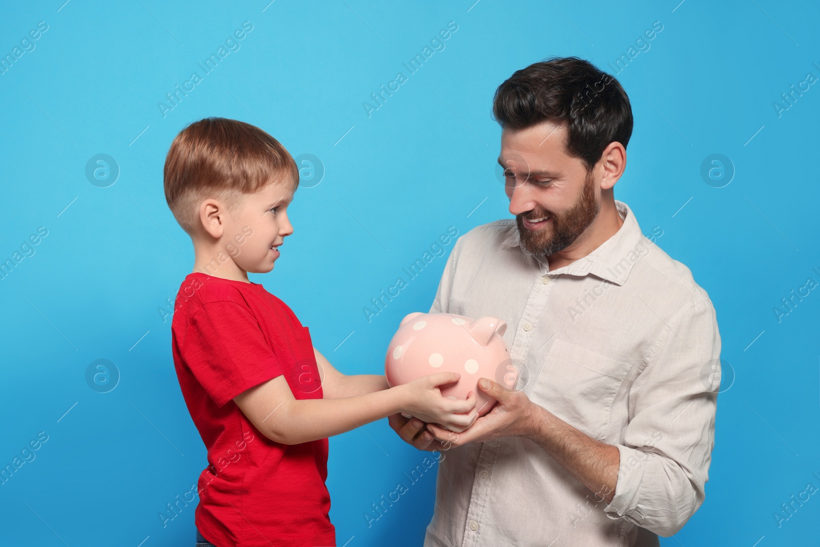 Photo of Father and his son with ceramic piggy bank on light blue background