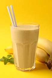 Photo of Glass of tasty banana smoothie with straws, fresh fruits and mint on yellow background
