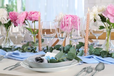 Photo of Beautiful table setting. Plate with greeting card, napkin and branch near glasses, peonies, burning candles and cutlery on table in room
