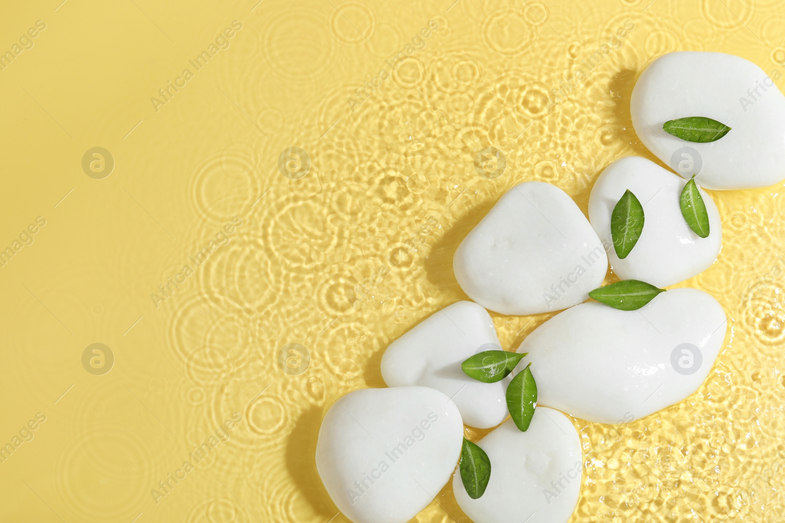 Photo of Spa stones and green leaves in water on yellow background, flat lay. Space for text