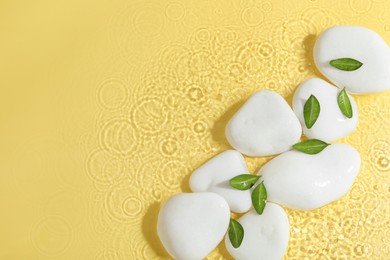 Spa stones and green leaves in water on yellow background, flat lay. Space for text