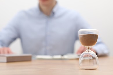 Photo of Hourglass with flowing sand on desk. Man using calculator indoors, selective focus