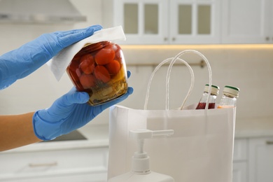 Photo of Woman cleaning newly purchased jar of pickled tomatoes with antiseptic wipe indoors, closeup. Preventive measures during COVID-19 pandemic