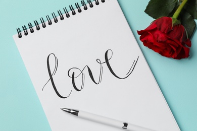 Photo of Notepad with handwritten word LOVE and rose on turquoise background, flat lay