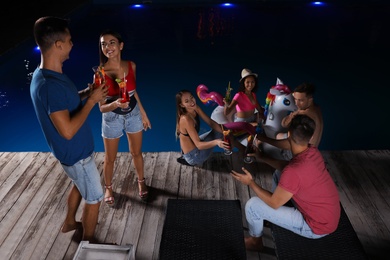 Photo of Happy young friends with refreshing cocktails relaxing near outdoor swimming pool at night