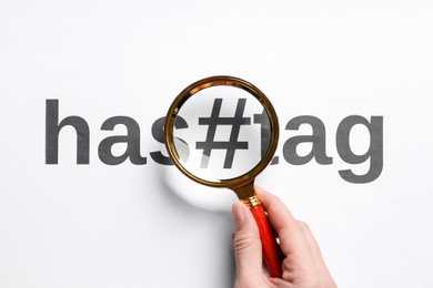 Photo of Woman holding magnifying glass over word Hashtag with symbol on white background, top view