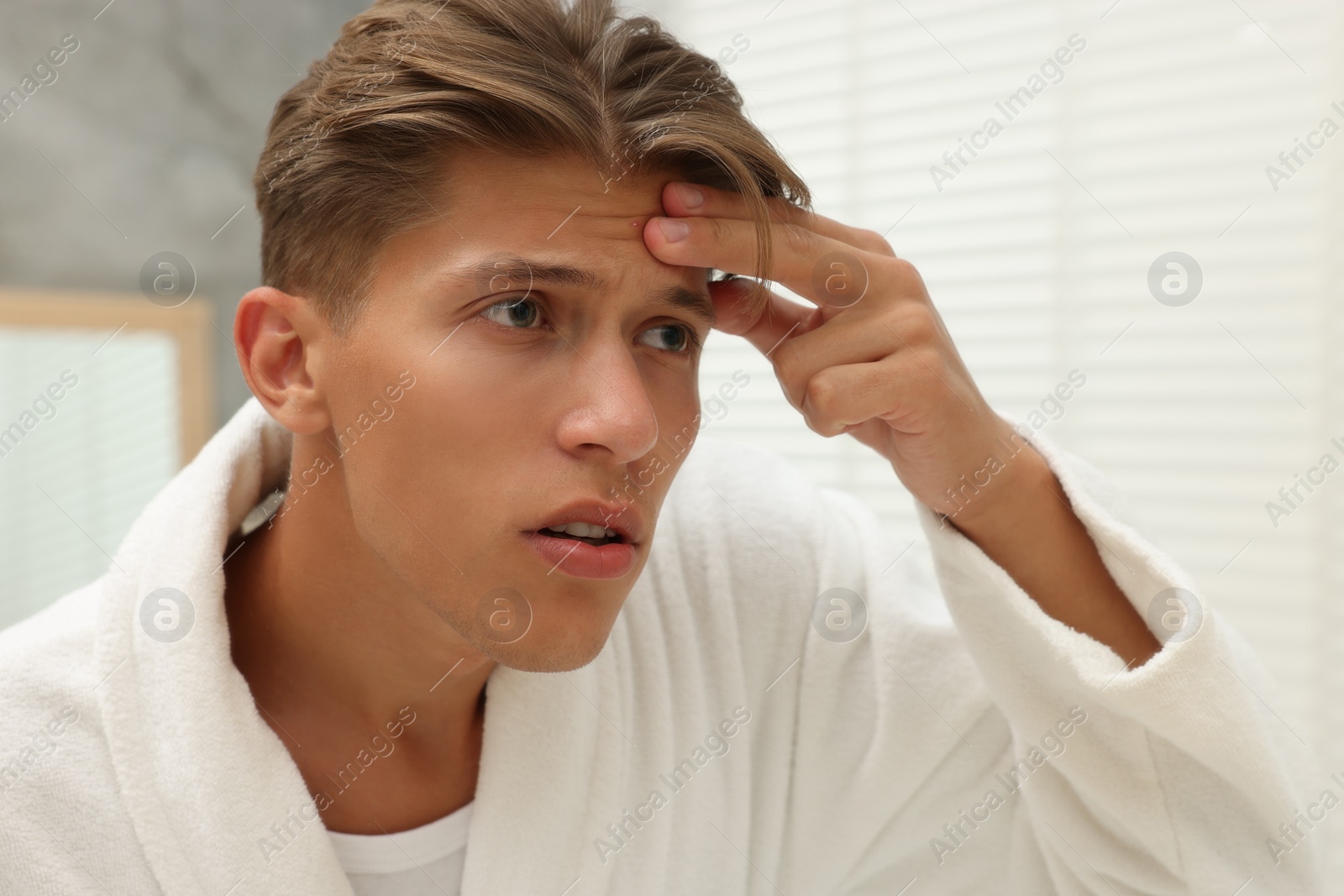 Photo of Upset young man looking at mirror and touching pimple on his face indoors. Acne problem