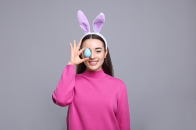 Photo of Happy woman in bunny ears headband holding painted Easter egg on grey background