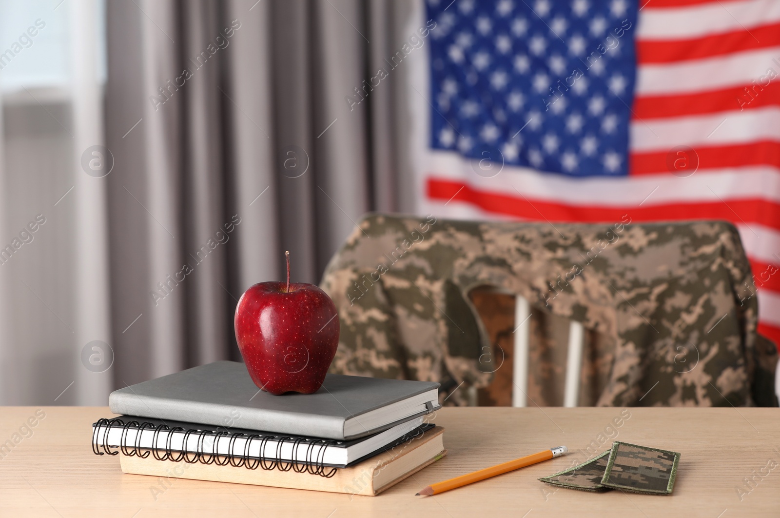 Photo of Notebooks, pencil, patches and apple on wooden table near flag of United States indoors. Military education