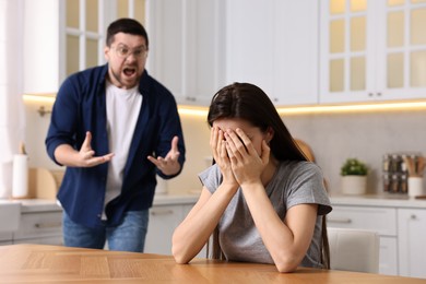 Stressed wife covering face with hands at table while her angry husband screaming at her in kitchen, selective focus. Relationship problems