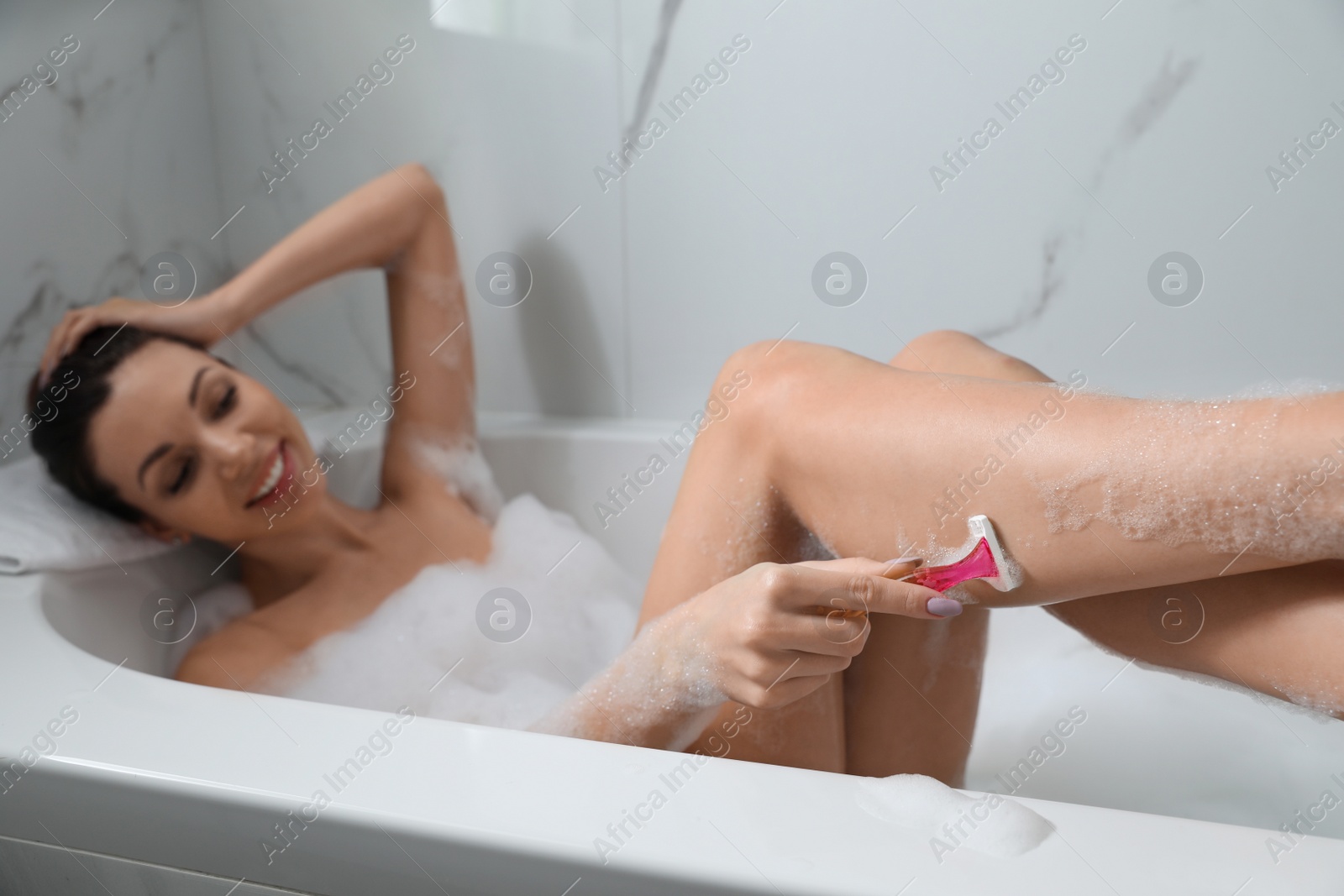 Photo of Young woman shaving legs while having bubble bath, focus on hand