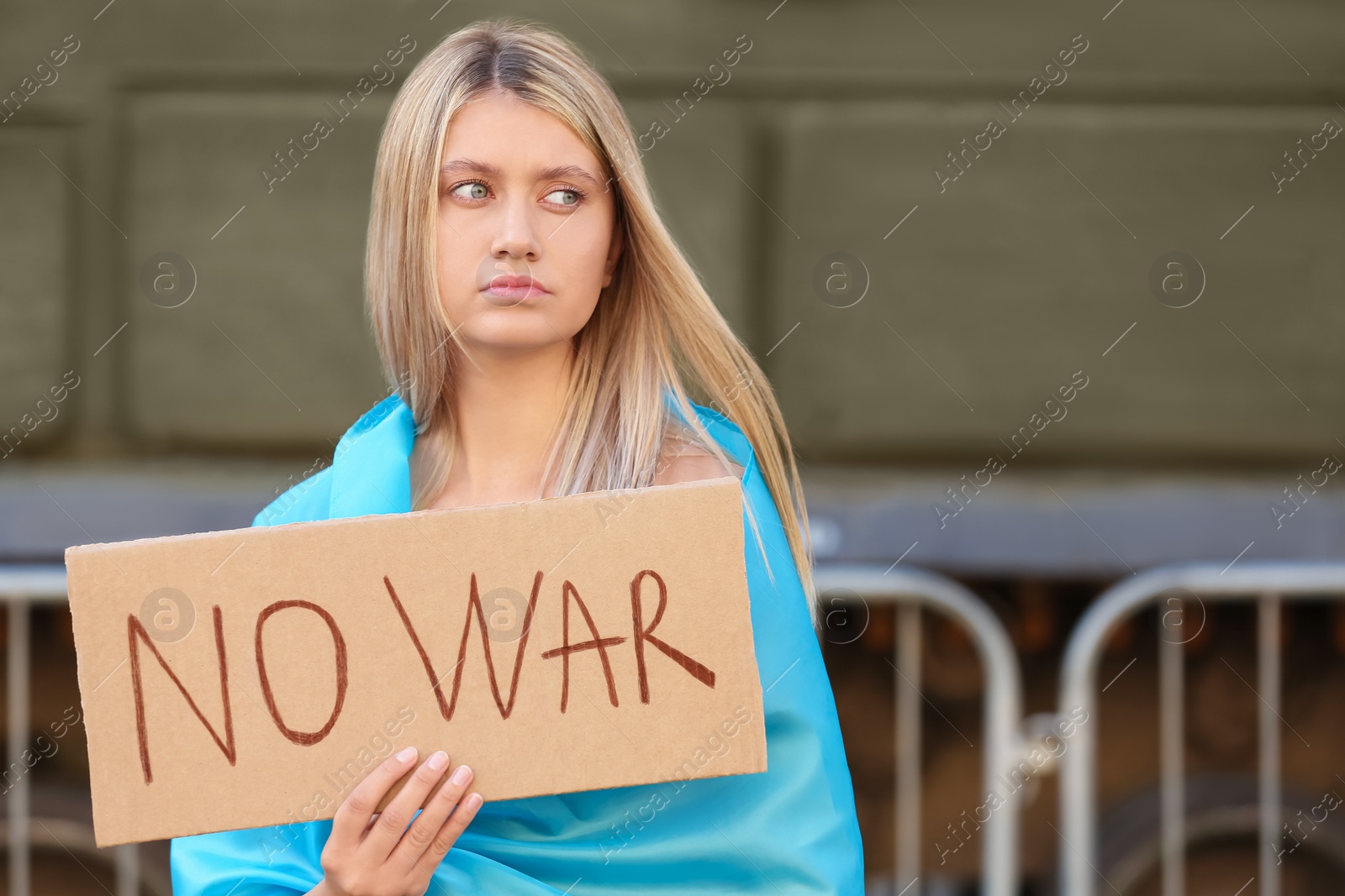 Photo of Sad woman wrapped in Ukrainian flag holding poster with words No War on city street