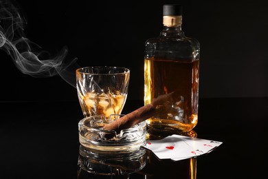 Photo of Smoldering cigar, ashtray, playing cards and whiskey on black mirror surface
