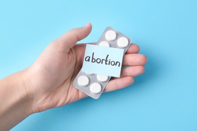 Photo of Woman holding paper note with word Abortion and pills on light blue background, top view