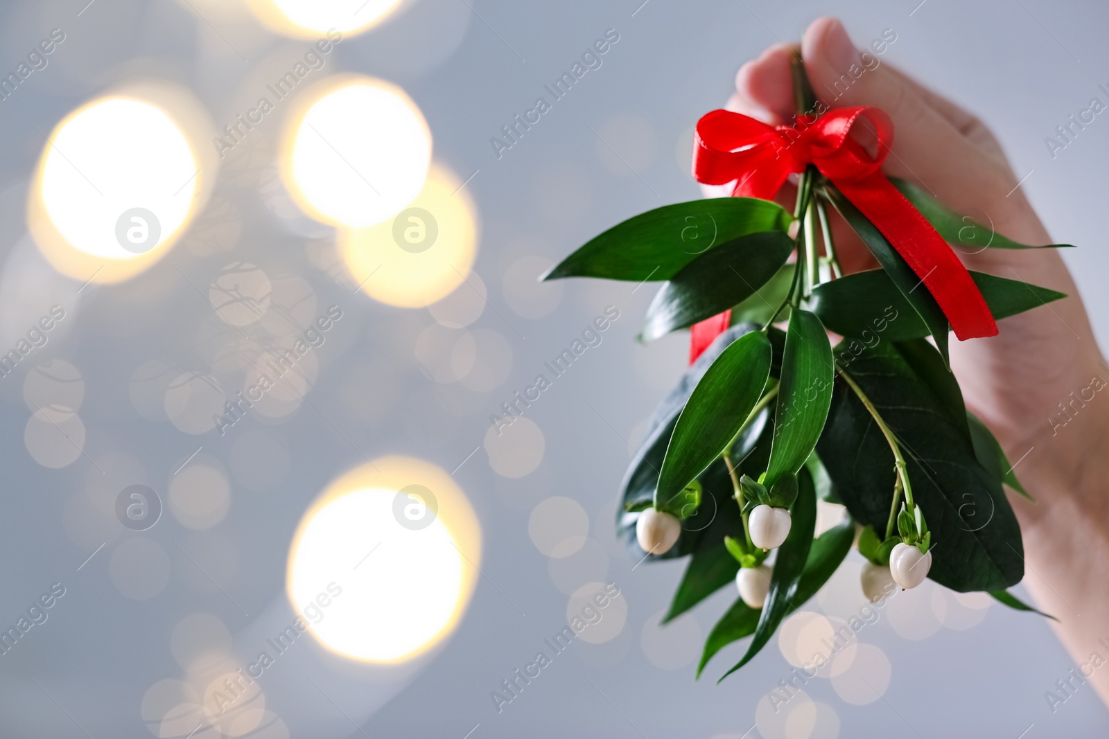 Photo of Woman holding mistletoe bunch with red bow against blurred festive lights, closeup and space for text. Traditional Christmas decor