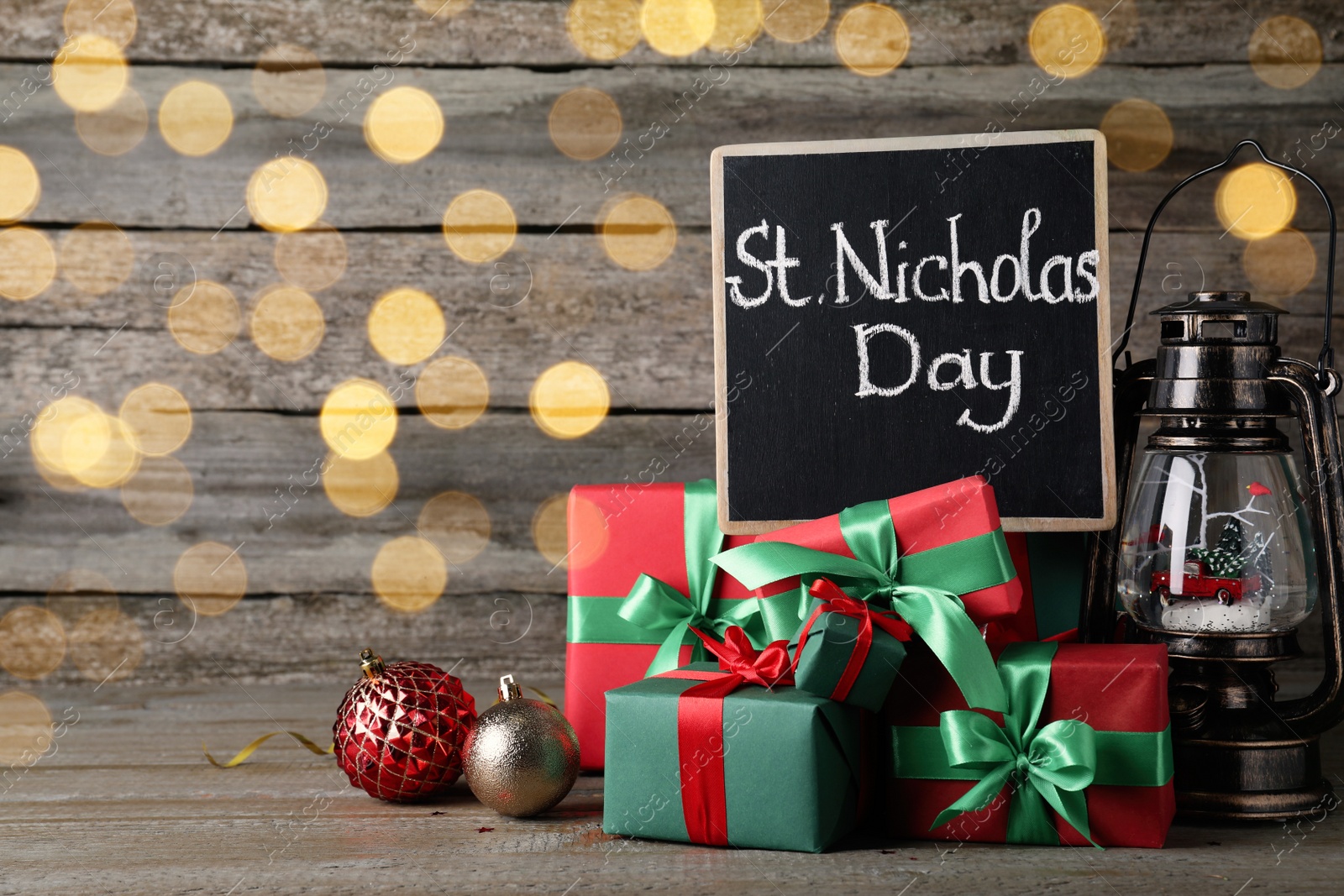 Photo of Blackboard with phrase St. Nicholas Day, gift boxes and festive decor on wooden table, space for text. Bokeh effect