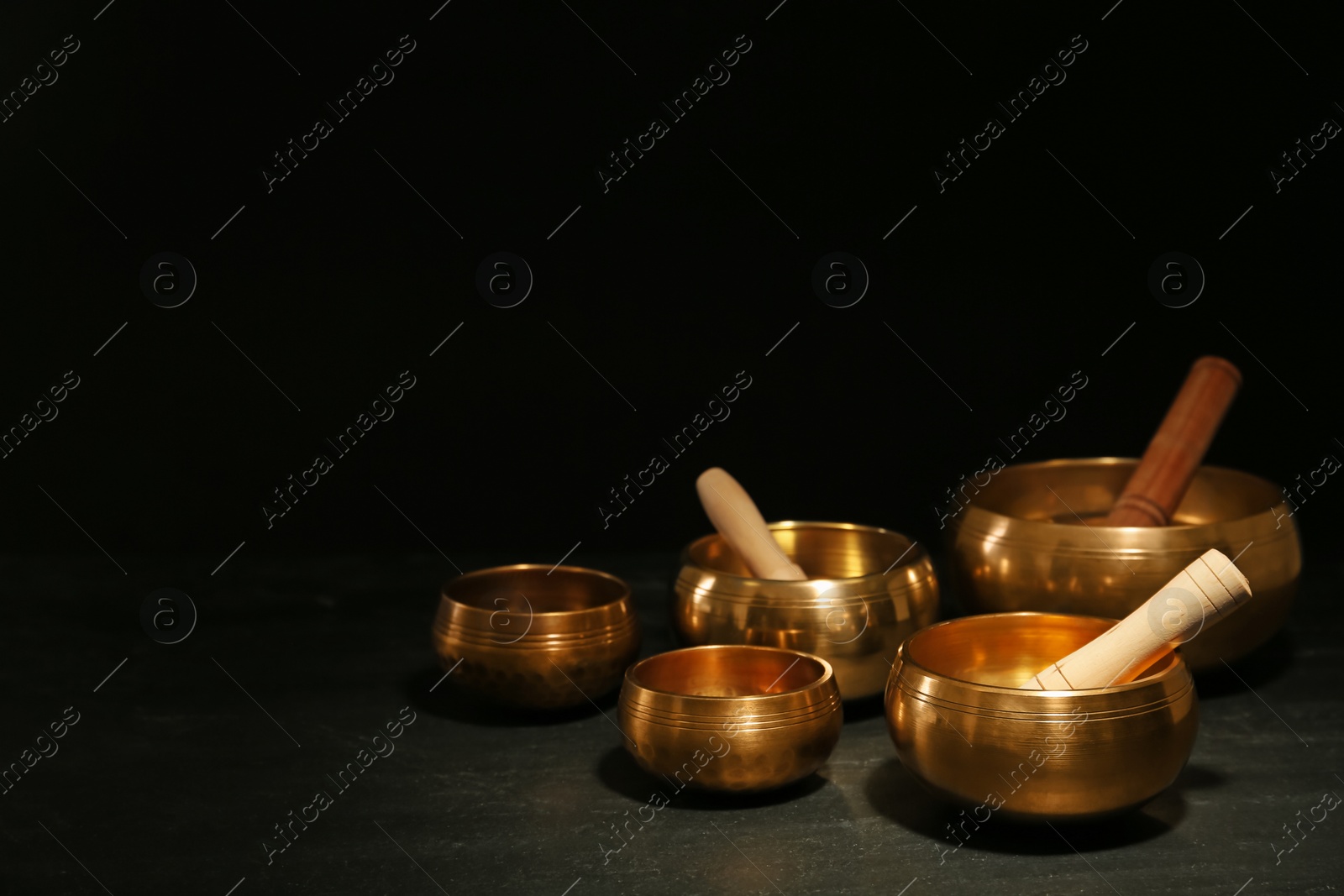 Photo of Golden singing bowls with mallets on black table against dark background, space for text