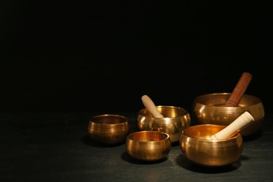 Photo of Golden singing bowls with mallets on black table against dark background, space for text