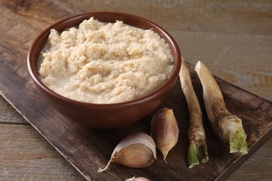 Spicy horseradish sauce in bowl, roots and garlic on wooden table, closeup