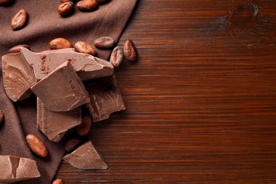 Photo of Pieces of tasty milk chocolate and cocoa beans on wooden table, flat lay. Space for text