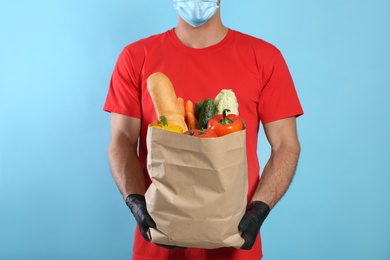 Photo of Courier in medical mask holding paper bag with food on light blue background, closeup. Delivery service during quarantine due to Covid-19 outbreak