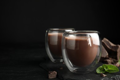 Glasses of delicious hot chocolate, chunks and fresh mint on black table. Space for text