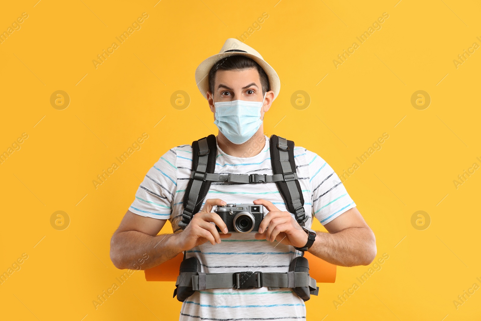 Photo of Male tourist in protective mask with travel backpack and camera on yellow background