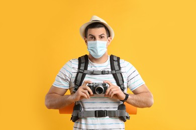Photo of Male tourist in protective mask with travel backpack and camera on yellow background