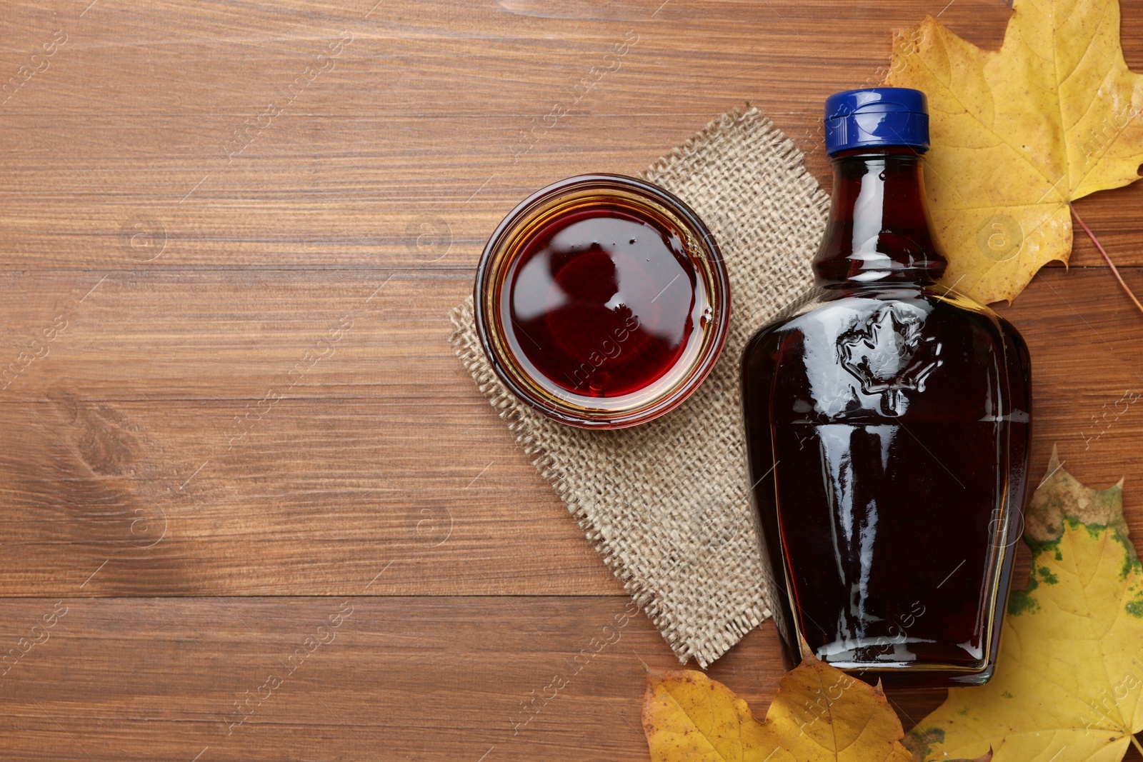 Photo of Bottle of tasty maple syrup, bowl and dry leaves on wooden table, flat lay. Space for text