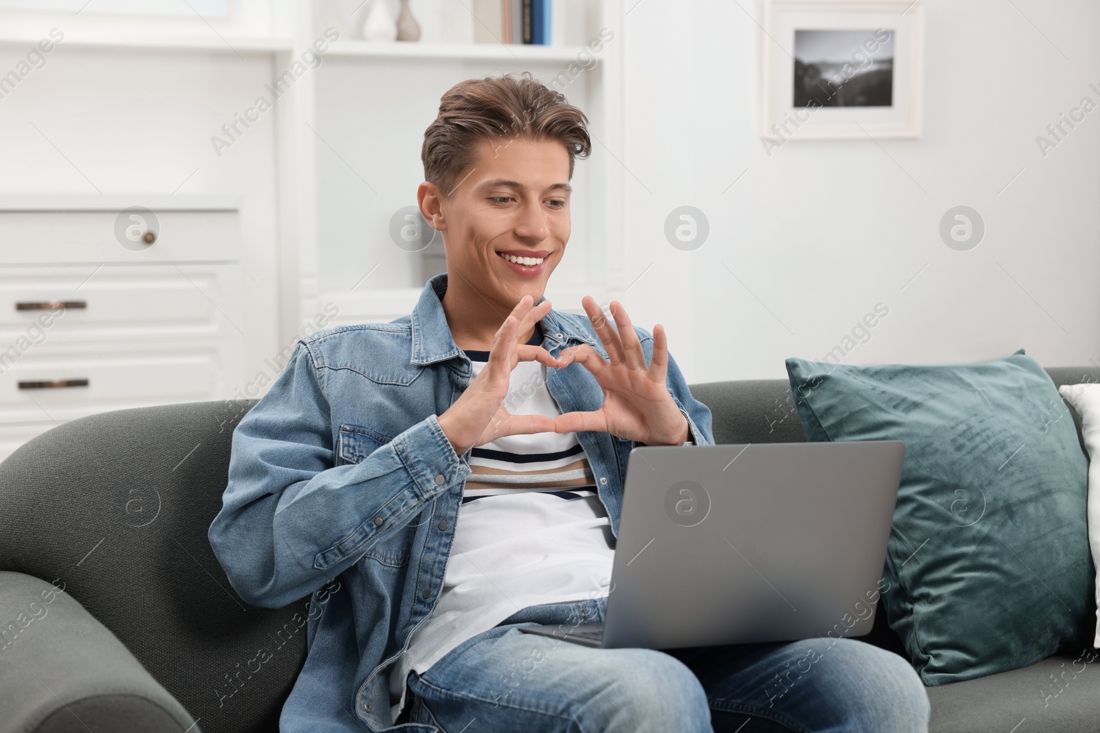 Photo of Happy young man having video chat via laptop and making heart with hands on sofa indoors. Long-distance relationship