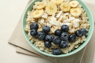 Tasty oatmeal with banana, blueberries, coconut flakes and honey served in bowl on beige table, closeup