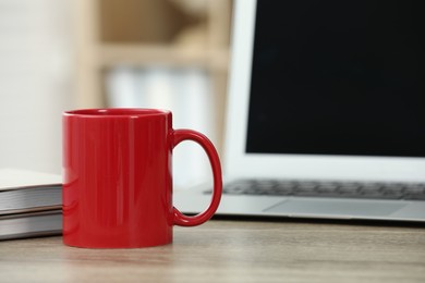 Photo of Red ceramic mug, notebooks and laptop on wooden table indoors. Space for text