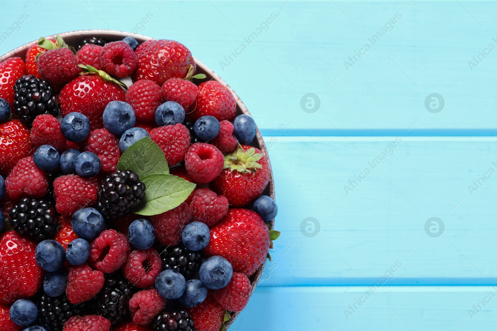 Photo of Different fresh ripe berries in bowl on light blue wooden table, top view. Space for text