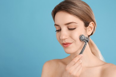 Young woman using metal face roller on light blue background, space for text