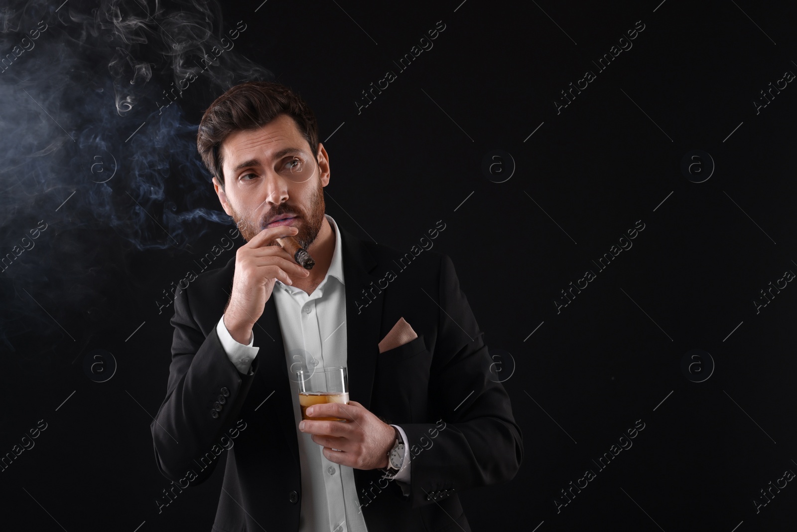 Photo of Handsome man in elegant suit with glass of whiskey smoking cigar on black background. Space for text