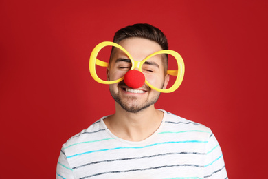 Photo of Happy young man with party glasses and clown nose on red background. April fool's day
