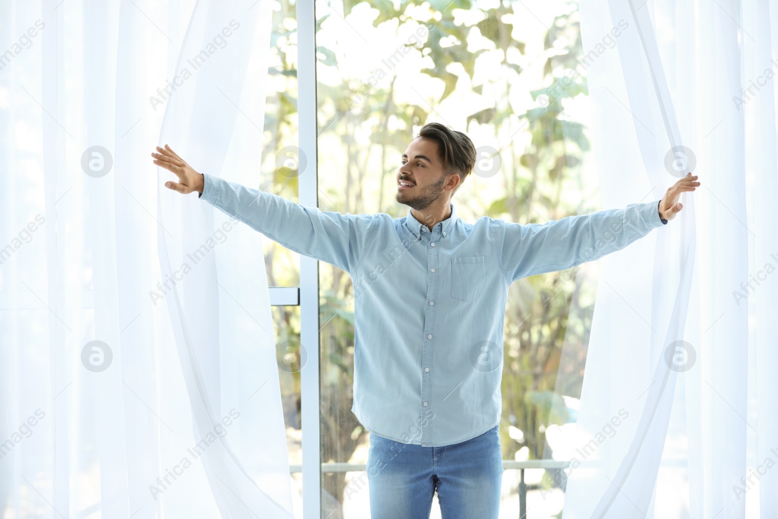 Photo of Young man opening window curtains at home