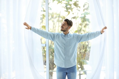 Young man opening window curtains at home