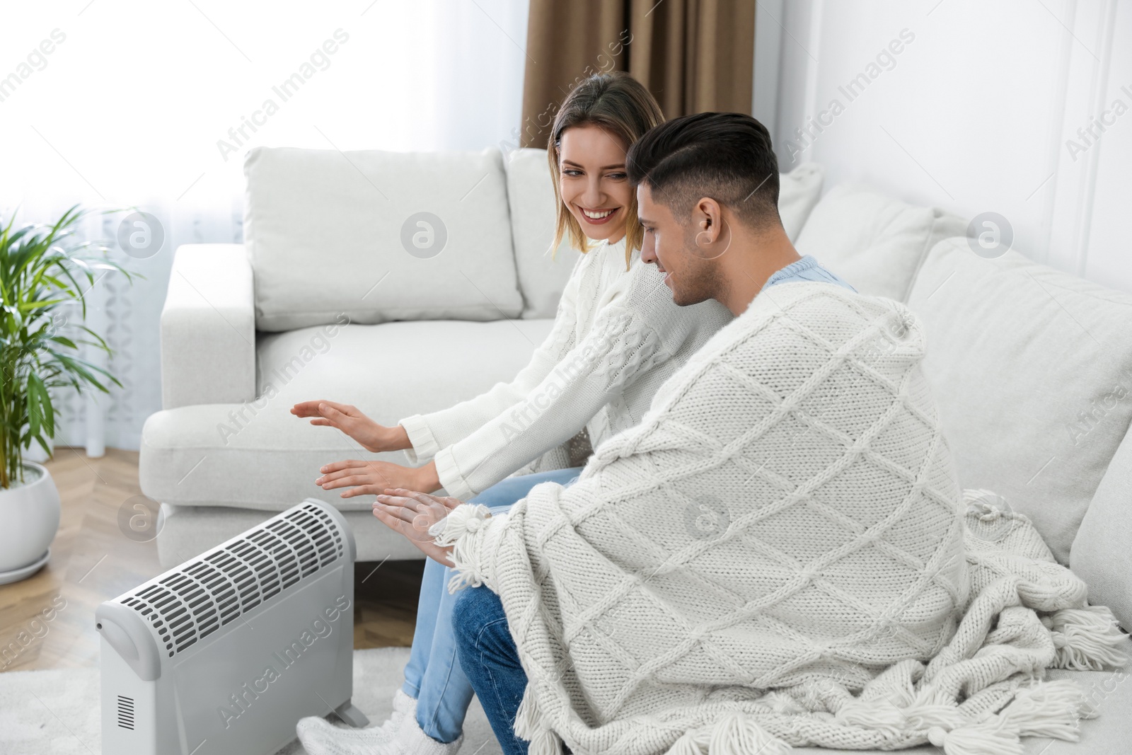 Photo of Happy couple sitting on sofa near electric heater at home