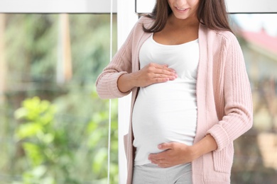 Photo of Pregnant woman standing near window at home, closeup