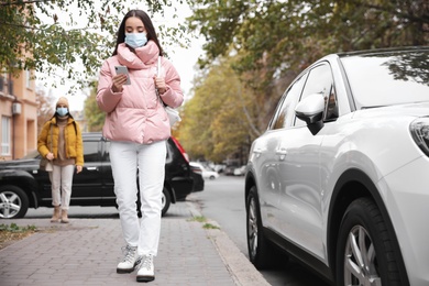 Photo of Young woman in medical face mask with smartphone walking outdoors. Personal protection during COVID-19 pandemic