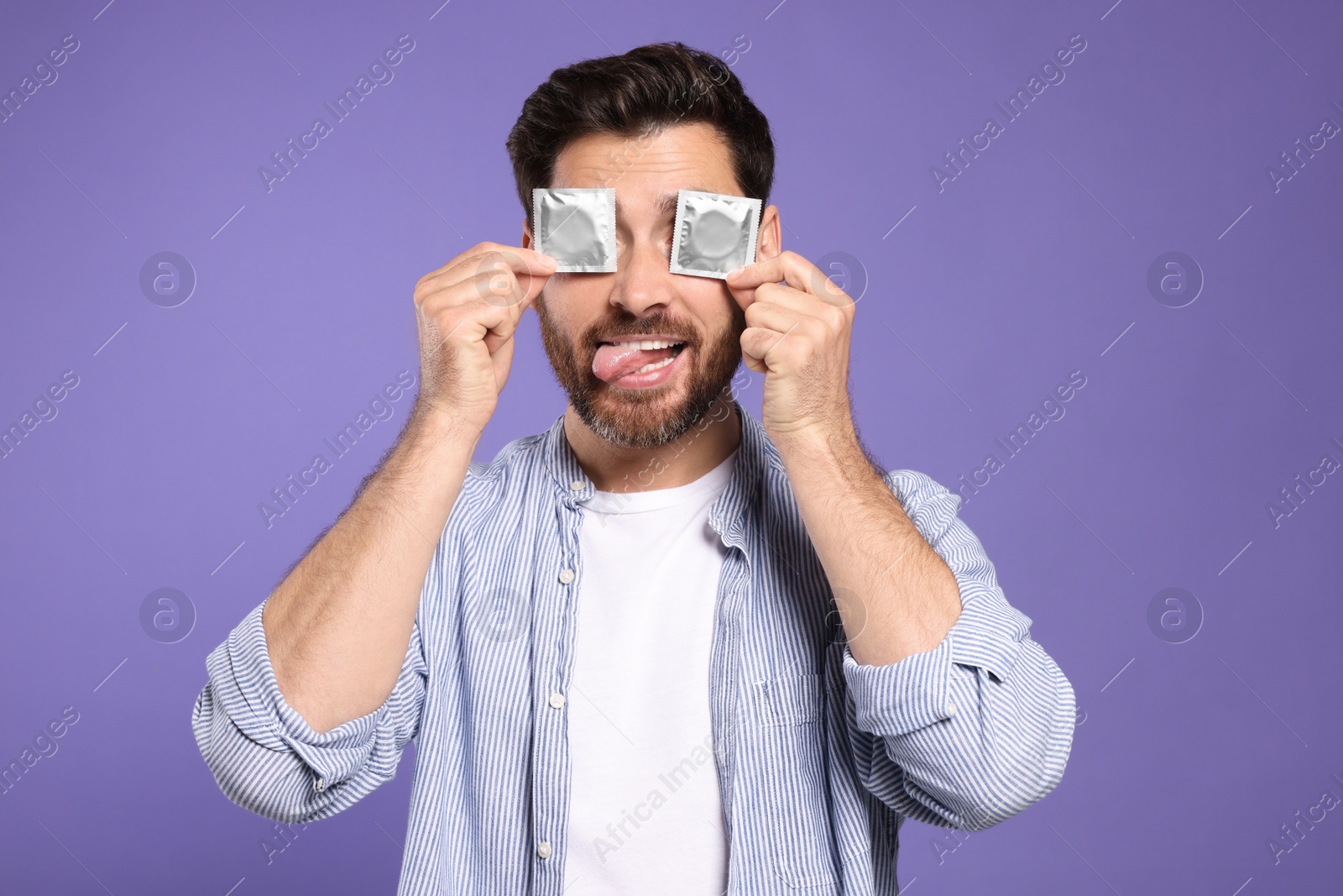 Photo of Man holding condoms near his eyes on purple background. Safe sex