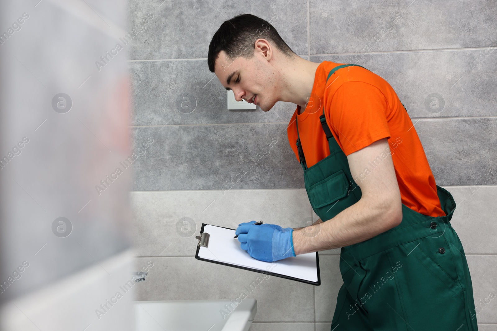 Photo of Young plumber writing results of examining sink in bathroom