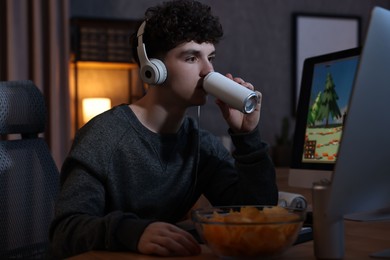 Photo of Young man with energy drink and headphones playing video game at wooden desk indoors