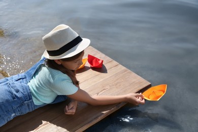 Photo of Cute little girl playing with paper boat on wooden pier near river