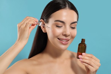 Beautiful young woman applying serum onto her face on light blue background